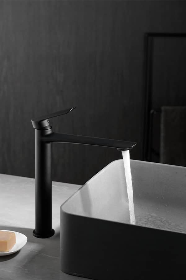 Robinet de lavabo noir mat de haute taille avec un design minimaliste, l'eau s'écoulant dans un lavabo rectangulaire assorti, sur fond de mur de salle de bain s