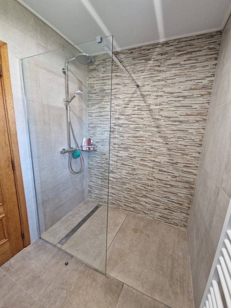 Salle de bain avec une grande douche vitrée, des carreaux de pierre naturelle sur le mur, un sol en carrelage beige et une serviette vert