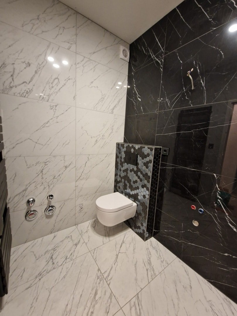 Vue d'une toilette moderne blanche à côté d'une paroi de douche en mosaïque grise et blanche, avec un sol en marbre blanc et des murs en
